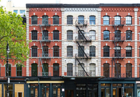 Red-brick building with fire escapes and storefronts, daytime.